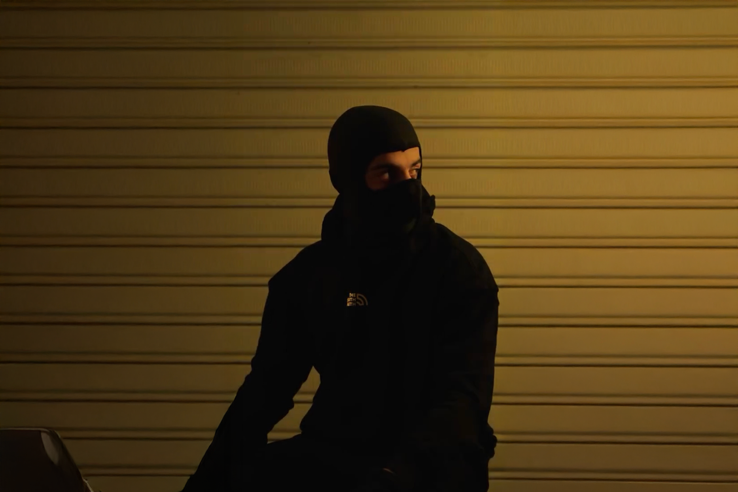 Person wearing a dark jacket and mask in front of a wall with yellow lights